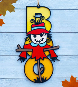 BOO wooden door sign featuring the letters B O O in a vertical format with various Halloween images pictured on top of the BOO. This one features a scarecrow.
