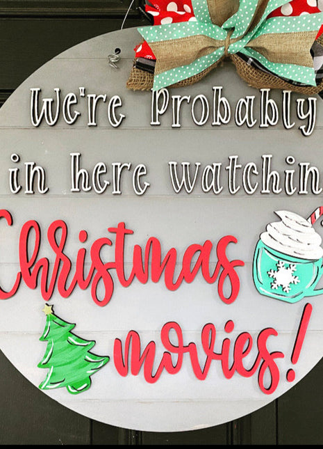 Round wooden door hanger that reads "We're probably in here watching Christmas movies!" Door hanger features also a fun ribbon, a christmas tree cutout, and a hot chocolate with whipped cream and peppermint cutout.