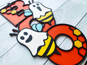 BOO wooden door sign featuring the letters B O O in a vertical format with various Halloween images pictured on top of the BOO. This one features two bumble bees dressed as ghosts. 