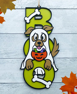 BOO wooden door sign featuring the letters B O O in a vertical format with various Halloween images pictured on top of the BOO. This one features a dog dressed as a ghost, holding a trick or treat pumpkin with candy in it. 