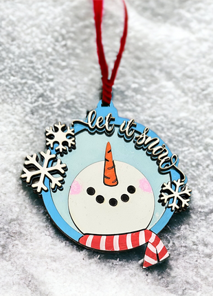 A wooden snowman ornament. Triple layer The snowman is looking up and has a carrot nose and a red and white striped scarf. There is a red ribbon. 