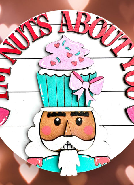 Round wooden door hanger that reads "I'm Nuts About You." The door hanger features a colorful nutcracker painted in pastel hues. Instead of a hat, he's wearing a cupcake on his head. 