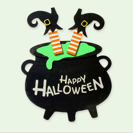 Wooden door hanger in the shape of a large black cauldron. A witch's legs and feet are upside down and sticking out of the cauldron. On the cauldron are words reading Happy Halloween. 