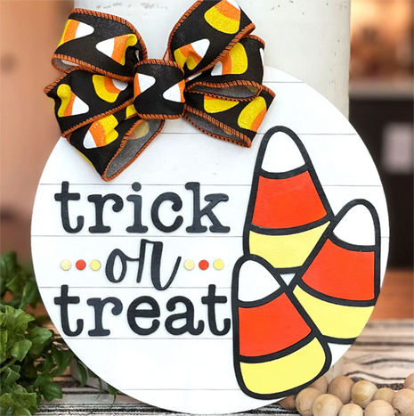 Round wooden door hanger with trick or treat written across it. The large candy corn candies are on the door hanger. There is also a large bow featuring candy corn. 