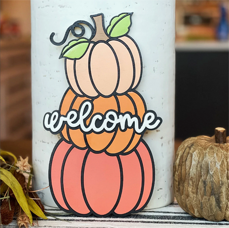 Wooden door hanger featuring three stacked pumpkins with the word welcome across it. The pumpkins are painted soft pastel colors. 