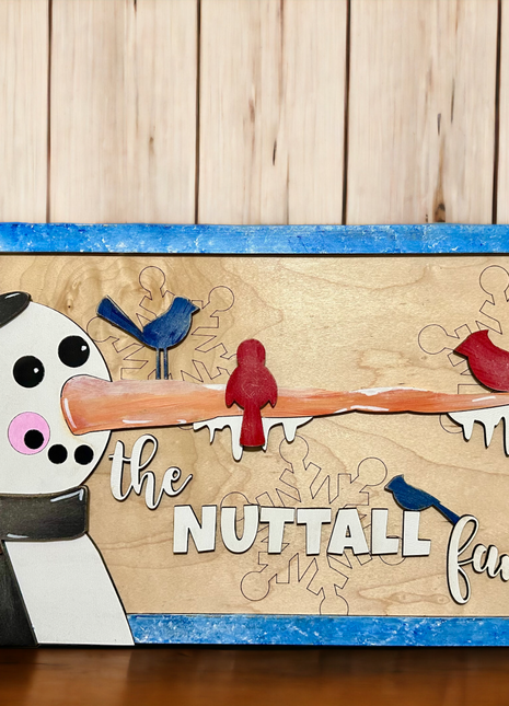 A horizontal wooden sign featuring a snowman with a carrot nose that's so long it spreads across the whole sign. Seated on the carrot and on the customized name "The Nuttall Family" are red and blue birds. The snowman is wearing a black top hat with snow on top of it and a black scarf with a red heart on it. 
