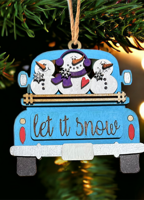 Blue wooden truck ornament with "let it snow" engraved on tailgate. There are three snowmen in the back of the truck smiling, with one of them wearing a purple scarf and ear muffs. 