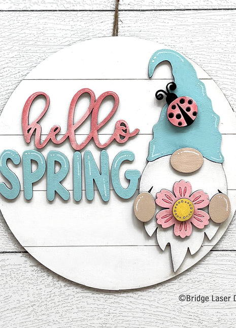 Round wooden door hanger that reads "hello spring." On the door hanger is a spring gnome with a light teal hat and he's holding a flower. There is a ladybug on his hat as well.