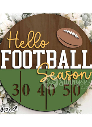 Round wooden door hanger "Hello Football Season." The bottom half features green to indicate grass and markings for the 30, 40 and 50 yard lines. There is a football at the top of the hanger.