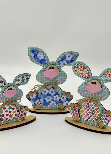 Wooden shelf sitter bunnies. Bunnies have big ears and are seated on a wooden base. While the bunnies are made of wood, they are covered with colorful scrapbook paper. 