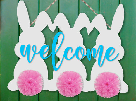 Wooden door hanger in the shape of three bunnies! Stretched across the back of the bunnies is a wooden cutout "welcome." The tails of the bunnies are poofy and pink! 